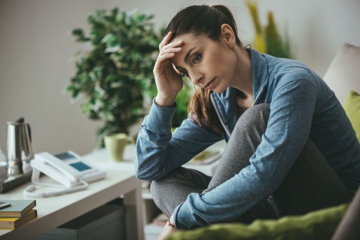 depressed woman sits on a sofa whilst clasping her head in her hand