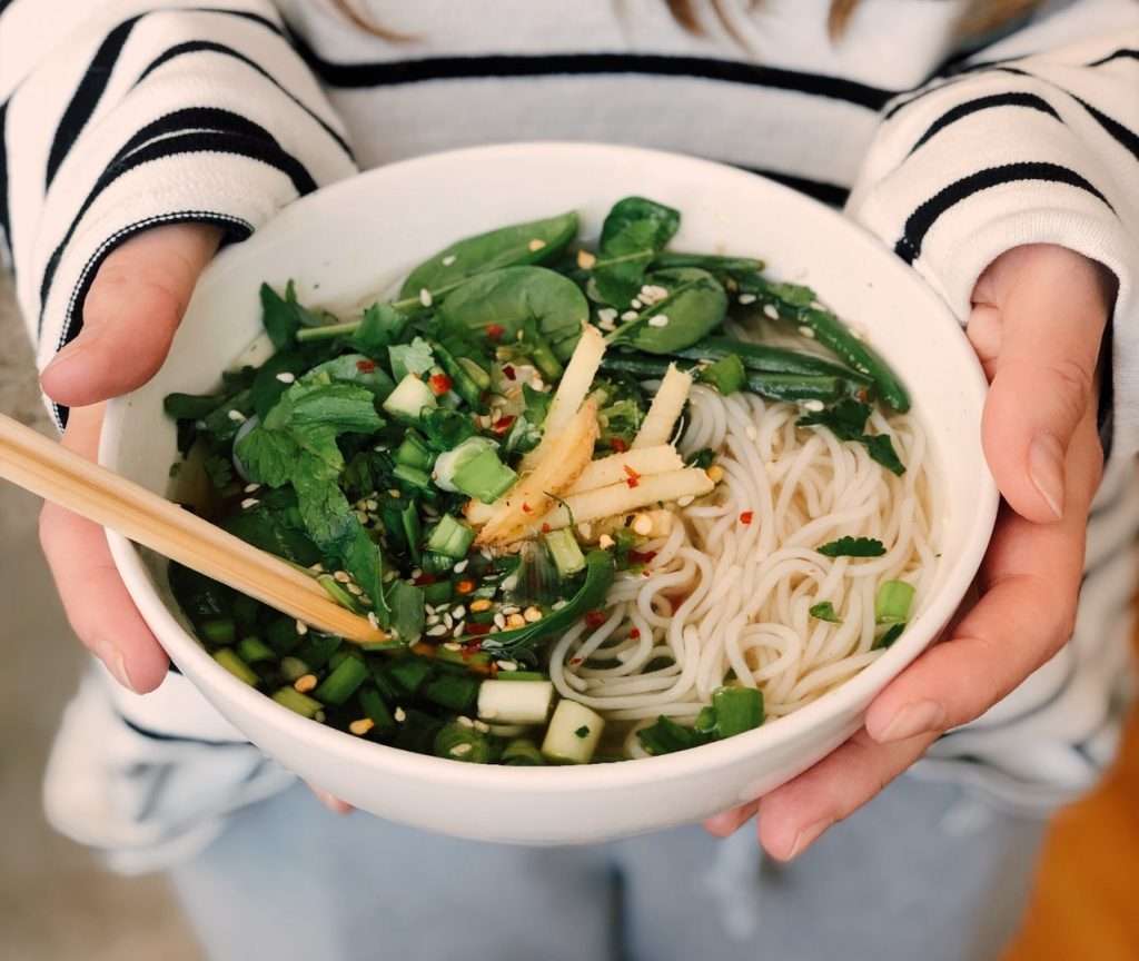 a female holding a bowl of chicken noodle soup with nutritious greens