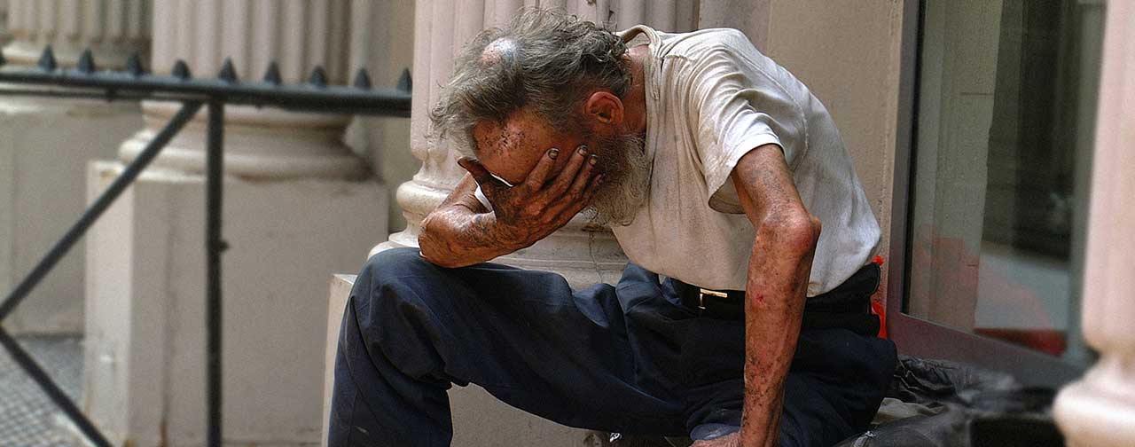 Elderly homeless man sitting on the street with hand covering his face, symbolizing poverty, addiction, and despair